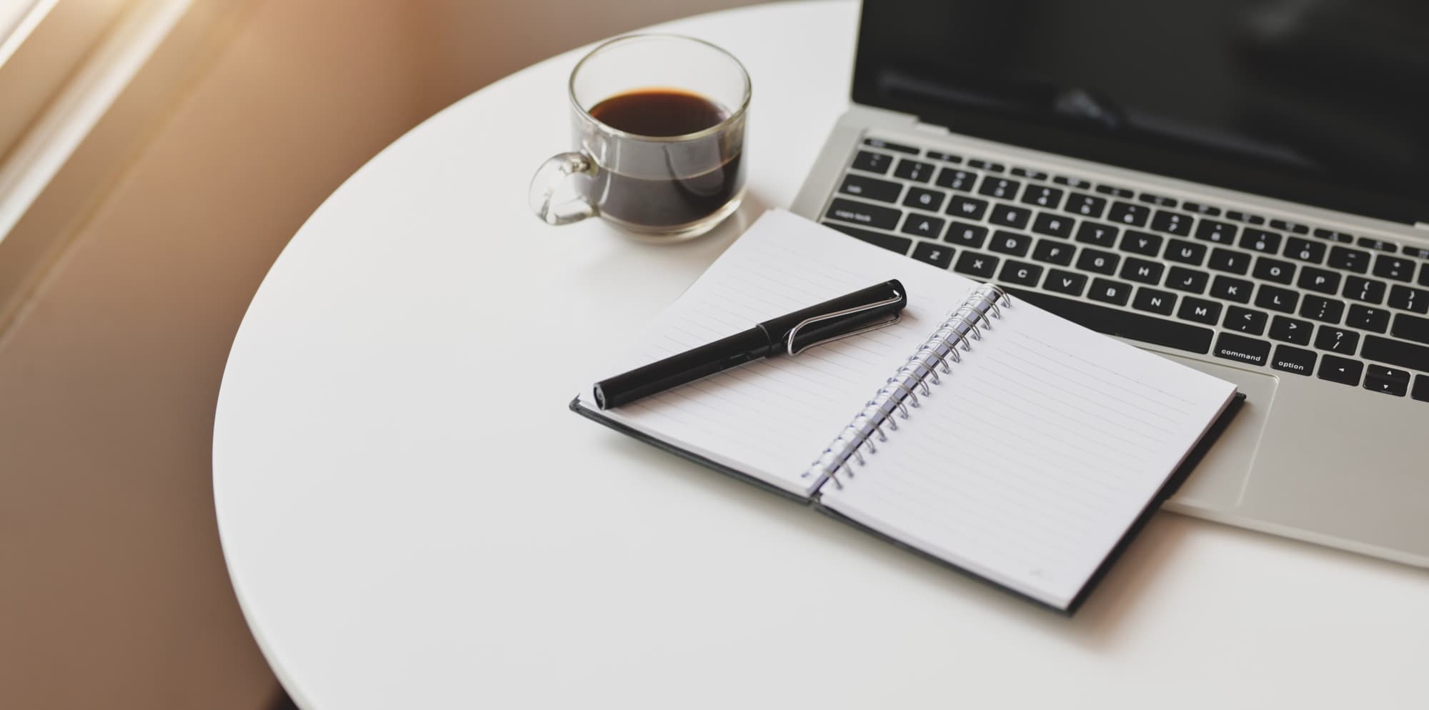 Laptop, open notebook with a pen, and a glass cup of black coffee on a round white table in warm morning light