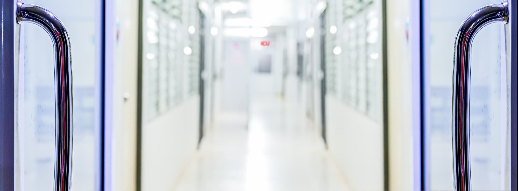 View through swing doors into a long empty hospital corridor with an illuminated ICU sign visible at the end of the hallway, evoking the operational reality behind a procurement decision